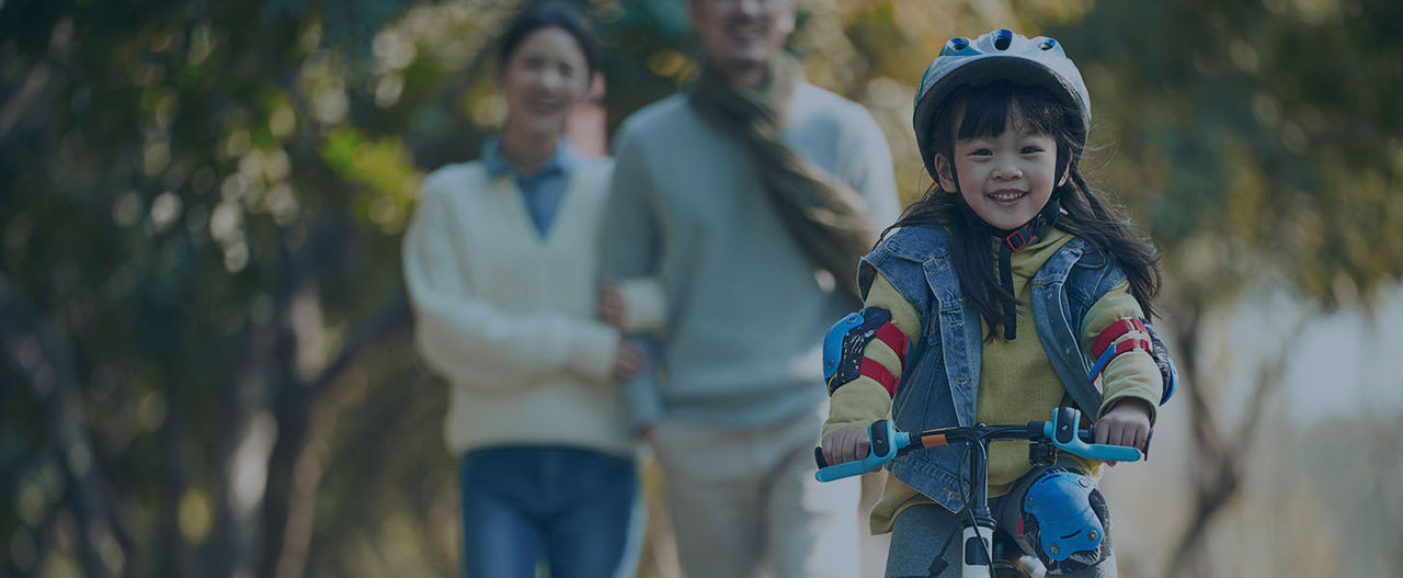 little asian girl with helmet and full protection gears riding bike in city park with parents watching from behind