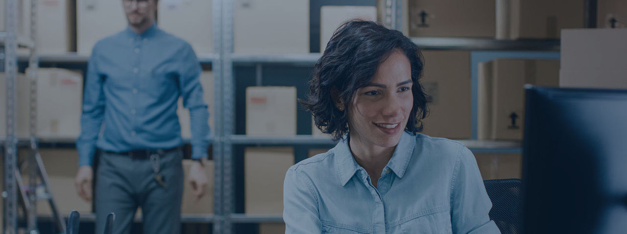 Female Inventory Manager Sitting at Her Desk and Using Personal Computer, Worker Puts Packages on the Designated Shelf. In the Background Rows of Cardboard Boxes with Products Ready For Shipment.
